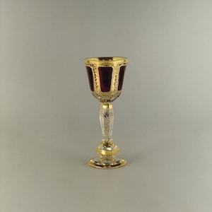 Puede incluir: Una copa de vidrio transparente con base roja y ribetes dorados. La copa tiene un patrón decorativo en el tallo y la base.