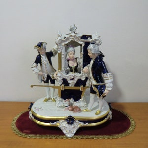 Può includere: Una statuetta in porcellana che raffigura una donna in una sedia a spalla portata da due uomini. La sedia a spalla è decorata con accenti dorati e le figure sono vestite di blu e bianco.