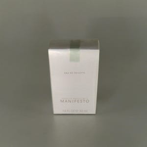 May include: A white rectangular perfume box with the text "EAU DE TOILETTE" and "MANIFESTO". The box is sealed with clear plastic and green tape. It contains 30 ml of perfume. The design is minimalist.