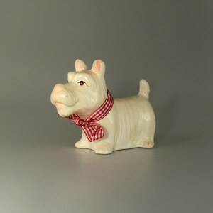 May include: White ceramic figurine of a dog wearing a red and white checkered bow. The dog is sitting with its head tilted to the side.