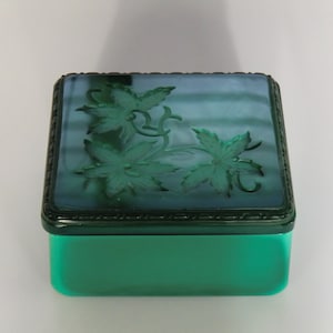 以下が含まれることがあります： 蓋に浮き彫りの花柄が施された正方形の緑色のガラス製の小物入れ。蓋は箱よりも濃い緑色です。