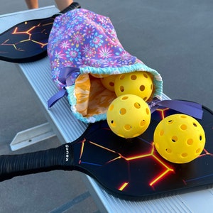 May include: Two black pickleball paddles with a glowing orange and red lava-like crack pattern and the brand name Gonex printed on the handle, accompanied by three bright yellow perforated pickleballs inside a purple floral drawstring bag.