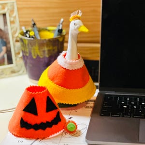 May include: A white goose figurine wearing a candy corn costume and a small hat. A felt pumpkin decoration with a jack-o'-lantern face sits nearby. A small green and orange decoration is also present. The items are Halloween-themed.