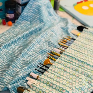 May include: A blue and white fabric roll with a green and white polka dot lining. The roll is filled with various paint brushes and pens.