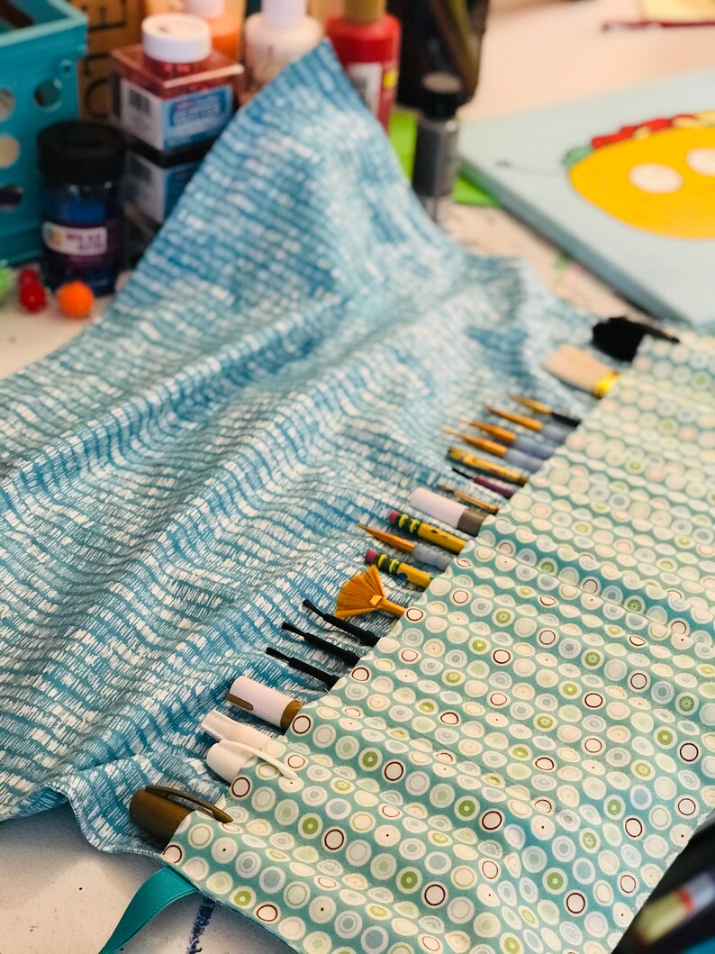 May include: A blue and white fabric roll with a green and white polka dot lining. The roll is filled with various paint brushes and pens.