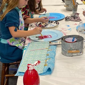 May include: Two children are painting ceramic plates with pink and blue paint. The plates are on a white table with a blue and white patterned fabric, a red paint bottle, a metal bucket with a red cup, and a water bottle.