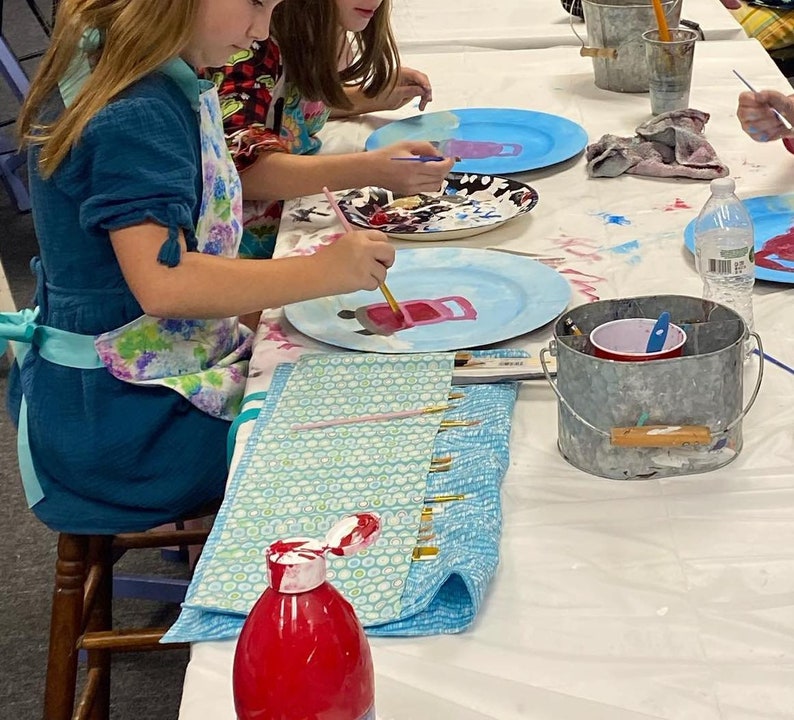 May include: Two children are painting ceramic plates with pink and blue paint. The plates are on a white table with a blue and white patterned fabric, a red paint bottle, a metal bucket with a red cup, and a water bottle.