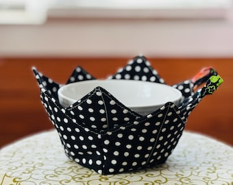 May include: A black and white polka dot bowl cozy with a white bowl inside. The cozy has a handle on each side and is sitting on a green and white patterned table cloth.