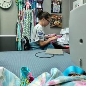 May include: A person is sitting at a desk using a sewing machine. There are fabric scraps and a laptop on the desk. The person is wearing a light blue shirt with a graphic on it.