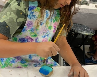 May include: A child paints a rock with blue, green, and white paint using a yellow paintbrush. The rock is sitting on a white surface.