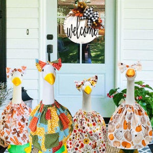 May include: Four white goose statues with yellow beaks and colorful fabric skirts and bow headbands. The skirts feature autumn-themed patterns with leaves, pumpkins, and floral designs. A welcome sign hangs on the door in the background.
