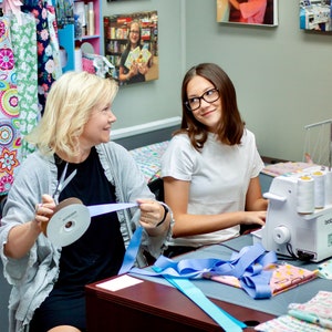 May include: Two people are working on a sewing project. One person is holding a spool of blue ribbon and a pair of scissors. The other person is using a sewing machine. The sewing machine is white with a black foot pedal and a black power cord. There are spools of thread on the sewing machine.
