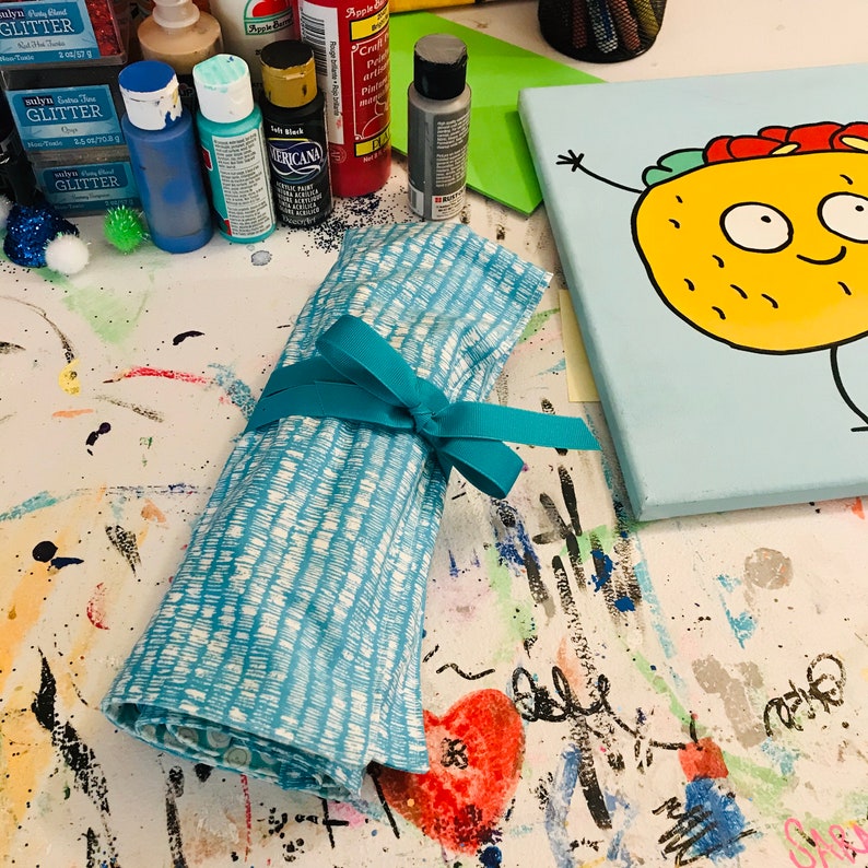 May include: A blue and white fabric roll tied with a turquoise ribbon. The fabric has a white stripe pattern. The roll is lying on a white surface with colorful paint splatters.