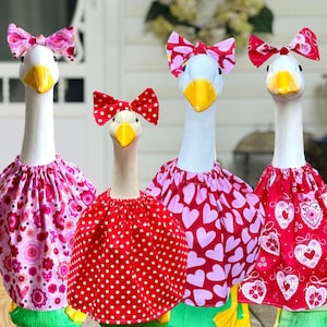 May include: Four white goose figurines wearing fabric outfits and matching bows. The outfits feature pink floral, red polka dot, pink heart, and red heart patterns. Each goose has a bright yellow beak and a white neck, standing in a row.