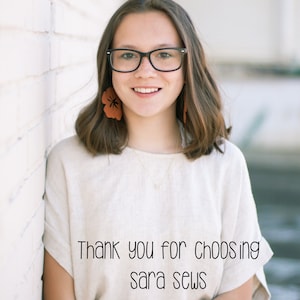 May include: A young woman with brown hair wearing glasses and a white shirt with the text "Thank you for choosing sara sews" on it. She is standing in front of a white brick wall.