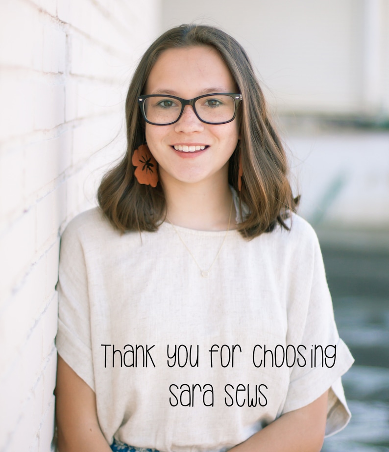 May include: A young woman with brown hair wearing glasses and a white shirt with the text "Thank you for choosing sara sews" on it. She is standing in front of a white brick wall.