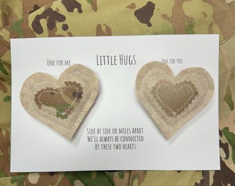 May include: Two fabric heart-shaped tokens attached to a white card with the text "LITTLE HUGS", "ONE FOR ME", "ONE FOR YOU", and "SIDE BY SIDE OR MILES APART WE'LL ALWAYS BE CONNECTED BY THESE TWO HEARTS". The hearts feature camouflage fabric.