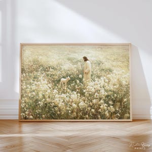 Puede incluir: Una impresión enmarcada representa una figura con una túnica blanca en un campo de flores blancas, con un cordero cerca. La obra de arte tiene una paleta de colores suave y cálida y está enmarcada en un marco de madera clara. La impresión está firmada "Rustic Berry Prints".
