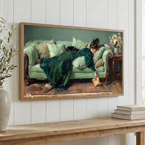May include: A framed print depicting a woman in a green dress on a green sofa. The artwork is in a classic painting style with a warm colour palette. A small black cat rests on the sofa. The print is displayed above a wooden table.