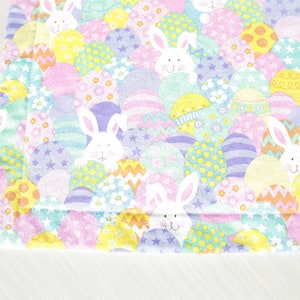 May include: A pastel pink, blue, and yellow fabric with a repeating pattern of Easter eggs and bunnies. The eggs are decorated with polka dots, stripes, and stars. The bunnies are white with pink noses and whiskers.