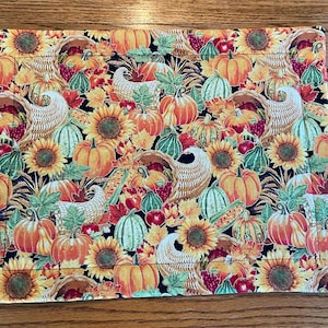 May include: A black fabric placemat with a Thanksgiving theme. The placemat features a repeating pattern of pumpkins, sunflowers, cornucopias, and fall leaves in shades of orange, yellow, green, and red.