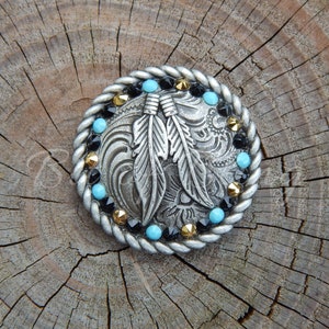 May include: A silver-toned belt buckle with a decorative design of two feathers and a swirling pattern. The buckle is surrounded by a rope-like border with turquoise and black rhinestones.