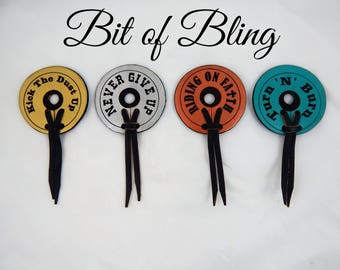 May include: Four round leather accessories with black cords. Each disc is a different color: gold, silver, orange, and teal. Each disc has a different phrase in black text: "Kick The Dust Up", "Never Give Up", "Riding On Faith", and "Turn 'N' Burn". The words "Bit of Bling" are above.