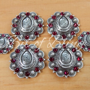May include: Set of six silver-toned horseshoe conchos with red rhinestone accents. The conchos are in two sizes, with the larger conchos having a more ornate design.