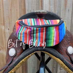 May include: A colourful striped saddle bag with a black zipper. The bag is sitting on a brown leather saddle.