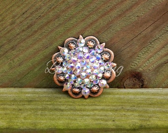 May include: A copper-colored, flower-shaped embellishment with a central cluster of iridescent rhinestones. The outer edge features a detailed, scalloped design. The rhinestones reflect a rainbow of colors, adding sparkle and visual interest to the piece.