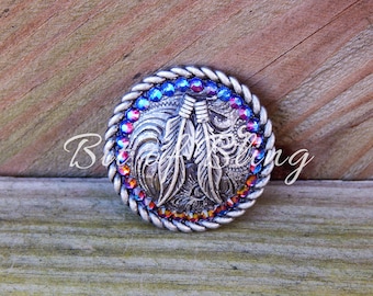 May include: A silver-toned, round button with a braided edge and colorful rhinestones. The center features a detailed design of feathers and decorative elements. The text "Blue Arting" is visible on the button.