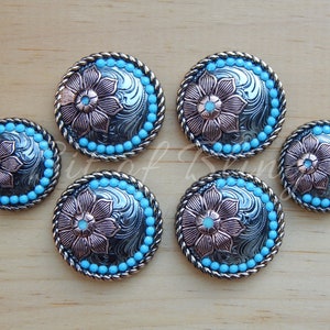 May include: A set of silver-coloured metal conchos with a floral design and turquoise accents. Each concho features a detailed flower in the centre, surrounded by a border of turquoise beads and a rope-like edge. The conchos are arranged on a wooden surface.