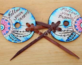 May include: Two round, hand-painted wooden hair accessories with the text "Follow your Dreams". Each disc features a blue and white background with a decorative feather and floral design, connected by brown leather straps.