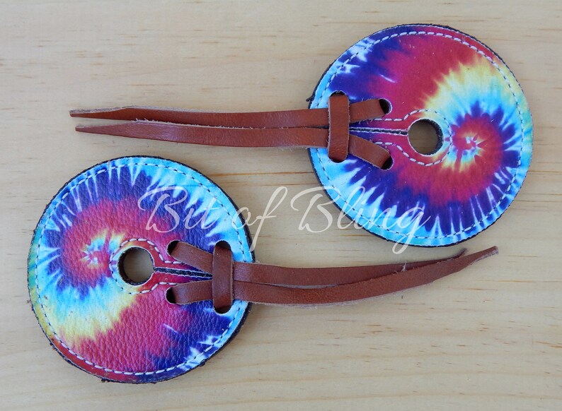 May include: Two tie-dye leather conchos with brown leather straps. The conchos have a swirl pattern in shades of blue, red, yellow, and white.