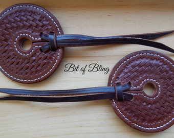 May include: Two brown leather spur straps with a woven design and white stitching. Each strap has a circular disc with a central hole and a long, pointed leather extension. The text "Bit of Bling" is visible.