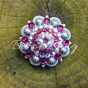 May include: A silver-toned, circular embellishment with a floral design. The center is filled with a cluster of pink and clear rhinestones. The text "Bit of bling" is written in cursive across the bottom.