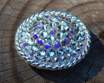 May include: A silver-toned belt buckle with a round, raised design. The buckle is covered in iridescent rhinestones, creating a sparkling effect.