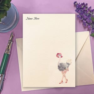 May include: Ivory stationery set with an ostrich illustration wearing a red hat and heels. The text "Name Here" is printed at the top. Includes a matching envelope. A pen and a small plant are on the purple surface.