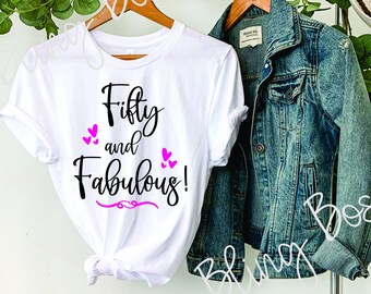 Puede incluir: Camiseta blanca con texto negro que dice "Fifty and Fabulous!" con corazones rosas. La camiseta está colgada en una percha de madera con una chaqueta vaquera y un sombrero de paja.