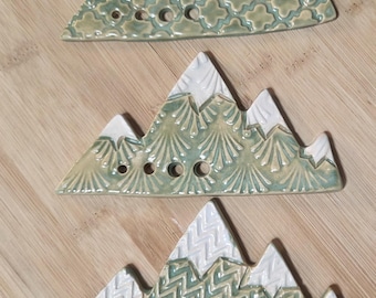 Puede incluir: Tres adornos de cerámica con forma de montaña, con picos esmaltados en blanco y bases de color verde salvia con distintos patrones en relieve. Cada pieza incluye cuatro orificios circulares en el borde inferior, con texturas de trébol, abanico y zigzag.