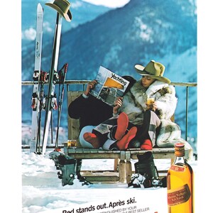 Op de afbeelding: Vintage advertentie voor Johnnie Walker Red Label whisky. De sc&egrave;ne toont een stel na het ski&euml;n, met ski's en laarzen. De tekst luidt "Red stands out. Apr&egrave;s ski" en de merknaam.