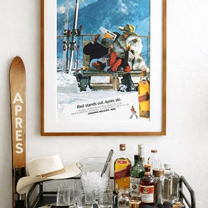 May include: A framed vintage advertisement with the text "Red stands out. Après ski." features a couple enjoying a drink after skiing. A bar cart with bottles, glasses, and a hat is in the foreground. A wooden ski is on the left.