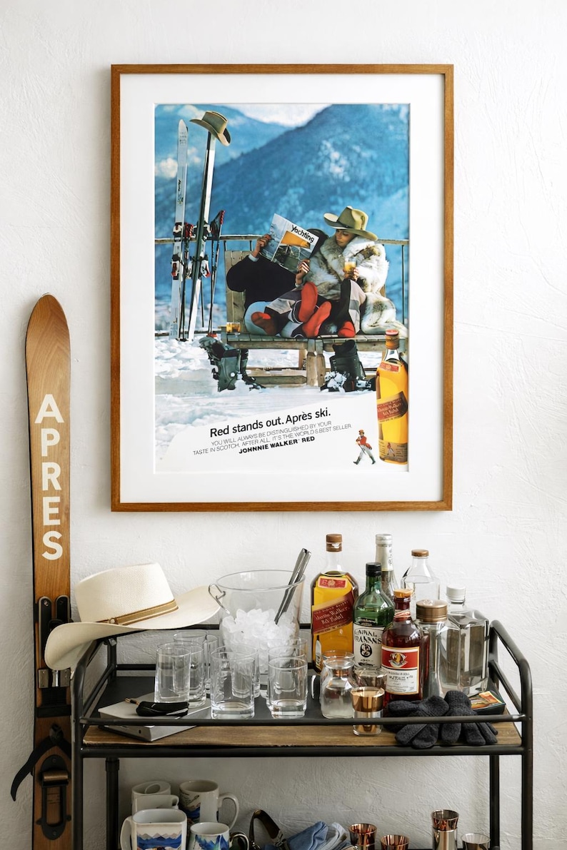 Op de afbeelding: Een ingelijste vintage advertentie met de tekst "Red stands out. Apr&egrave;s ski." toont een stel dat na het ski&euml;n van een drankje geniet. Een bartrol met flessen, glazen en een hoed staat op de voorgrond. Een houten ski staat links.