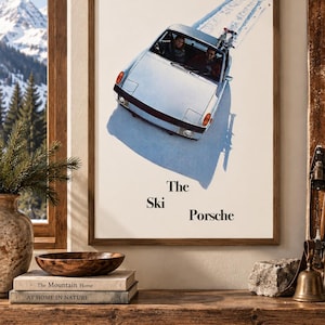 May include: A framed vertical wall art print featuring a silver convertible sports car driving on a snow-covered surface. The car carries skis on the back. The text "The Ski Porsche" is printed in black below the car on a white background.