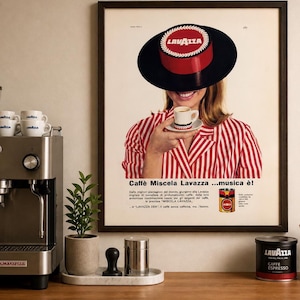 May include: A framed vintage Lavazza coffee advertisement. The image features a woman holding a cup of coffee, wearing a hat with the Lavazza logo. The text "Caffè Miscela Lavazza...musica è!" is displayed.