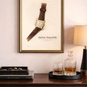 May include: A framed print of a Patek Philippe watch with a brown leather strap. The watch has a gold square face. The print is in a gold frame. A decanter, two glasses, and a tray are on a dark wood surface.