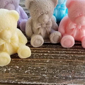 May include: A collection of pastel-colored teddy bear soaps. The bears are in shades of yellow, lavender, gray, blue, and pink. Each bear has a bow and is sitting on a wooden surface. These soaps are ideal for baby showers or as a gift.