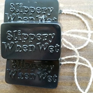 May include: Three black soap bars stacked on top of each other. Each bar has the words "Slippery When Wet" embossed on the front. The bars are tied together with a piece of twine.