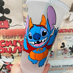 May include: A frosted plastic cold cup with a clear lid and a dark green straw. The cup features a graphic of the blue character Stitch wearing an orange pumpkin costume with a red cape and orange horns, smiling with visible teeth.