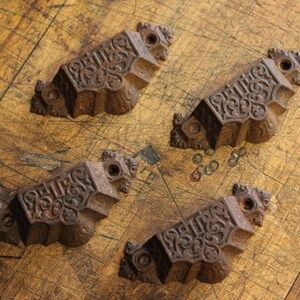 May include: Four antique bronze drawer pulls with ornate scrollwork and a weathered finish. The pulls are rectangular with a curved bottom and feature mounting holes on each end. These decorative hardware pieces are ideal for vintage furniture restoration.